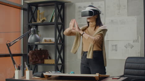 Young-female-engineer-in-office-in-virtual-reality-helmet-with-hands-makes-movements-imitating-the-work-of-graphic-interface.